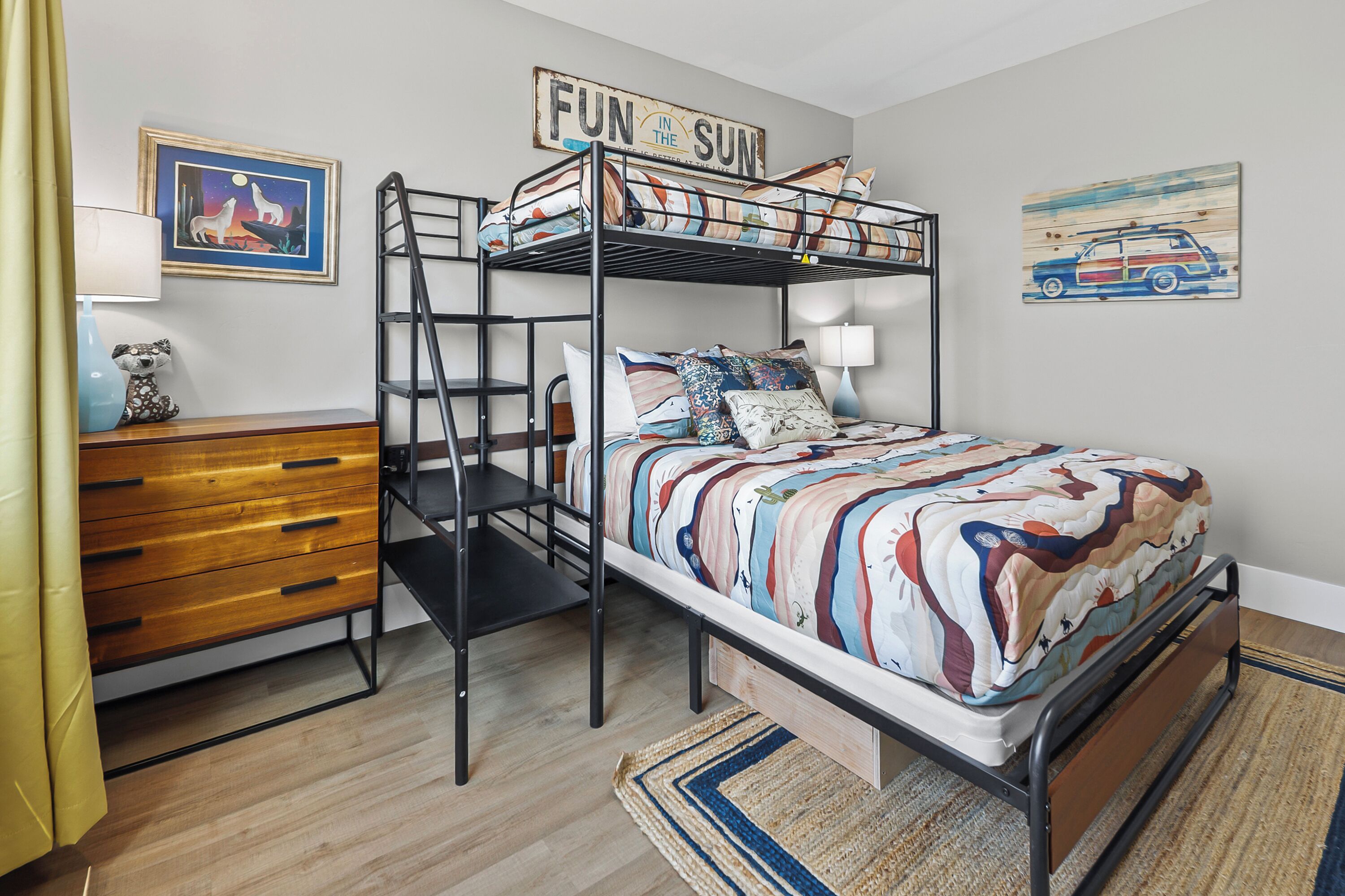 Un lit superposé avec une couvre-lit aux couleurs vives, une commode en bois, un bureau avec une lampe et des œuvres d’art accrochées au mur.