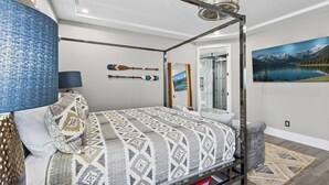 A four-poster bed with a patterned bedspread, a blue textured wall, and a painting of a lake. - New on VRBO! New Construction, Stylish Mega RV Garage House (Lake Havasu City)