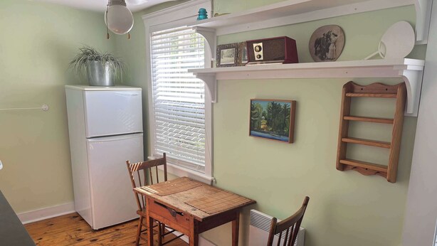 A compact kitchen with a dining table, chairs, a refrigerator, and a window with blinds. - Pentz Howe House Apartment, (Charlottetown)