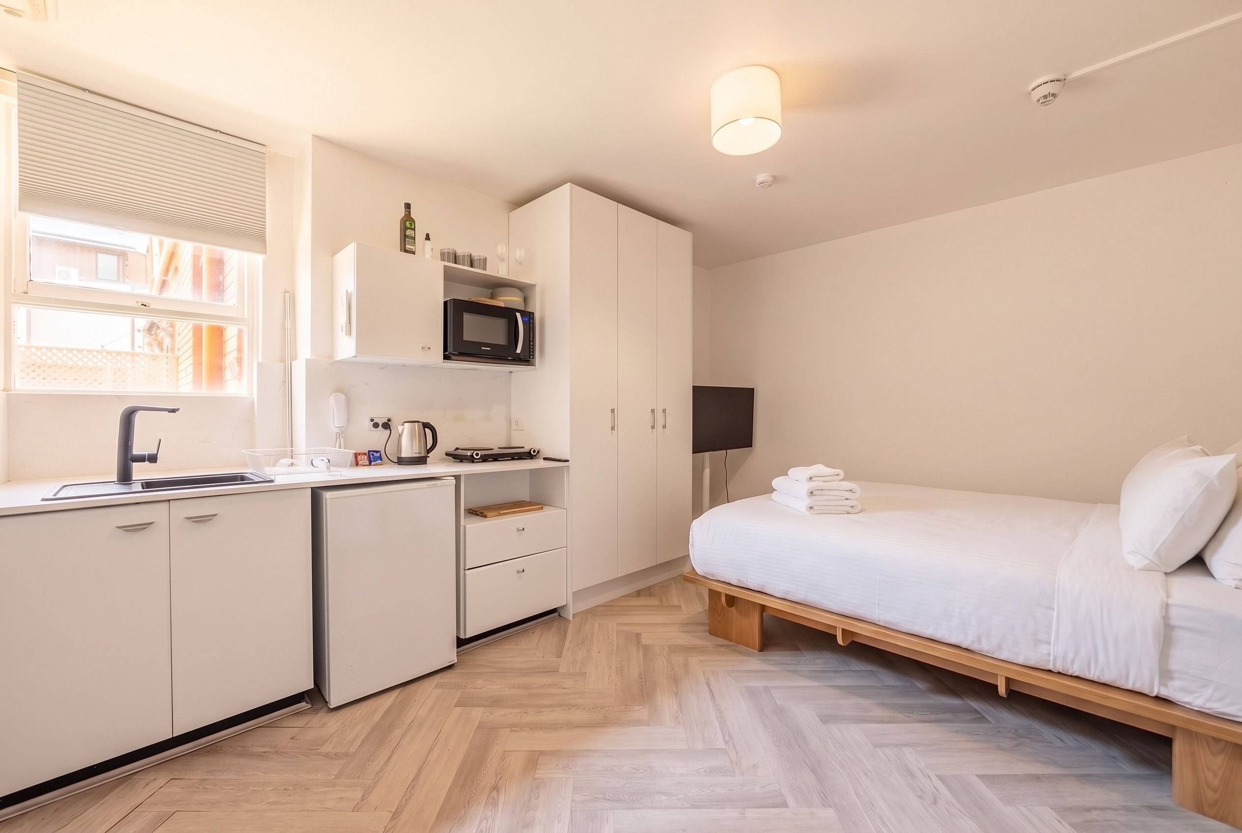 Una camera d'albergo compatta con angolo cottura, un letto con lenzuola bianche e pavimento in legno.