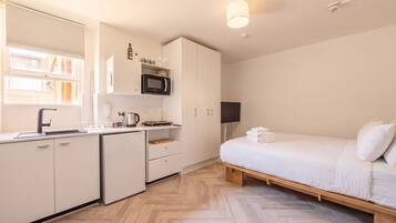 A compact hotel room with a kitchenette, a bed with white linens, and a wooden floor.