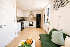 A compact living space with a kitchenette, a washing machine, a dining area with a fruit bowl, and a green sofa. - Cardiff 3-Bed Flat | Equipped Kitchen, Smart TV & Street Parking (Cardiff)