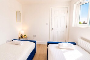 A hotel room with two beds, a white door, a window with a view, and a bedside table with a lamp. - Cardiff 3-Bed Flat | Equipped Kitchen, Smart TV & Street Parking (Cardiff)