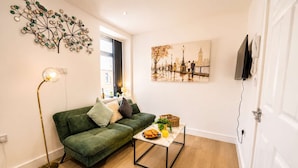A living room with a green sofa, a coffee table with breakfast items, a floor lamp, a wall art of a cityscape, and a flat-screen TV mounted on the wall. - Spacious 3BR Flat Sleeps 10 for Groups & Family (Cardiff)