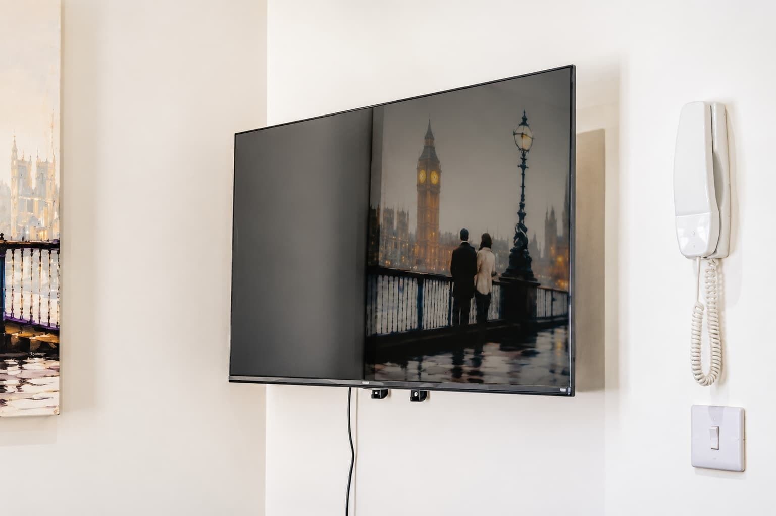 Seinälle asennettu litteänäyttöinen televisio, sen lähellä valkoinen puhelin ja valokatkaisin.