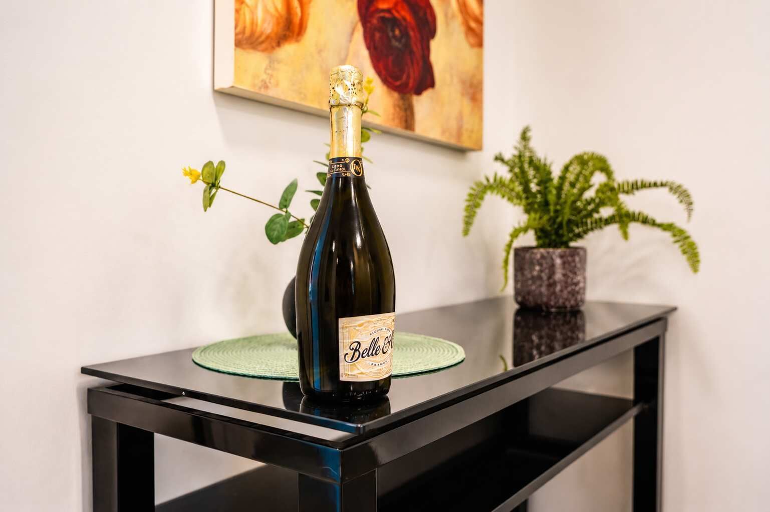 Une bouteille de parfum Belle posée sur une table en verre, avec une nappe verte, une plante en pot et un tableau en arrière-plan.