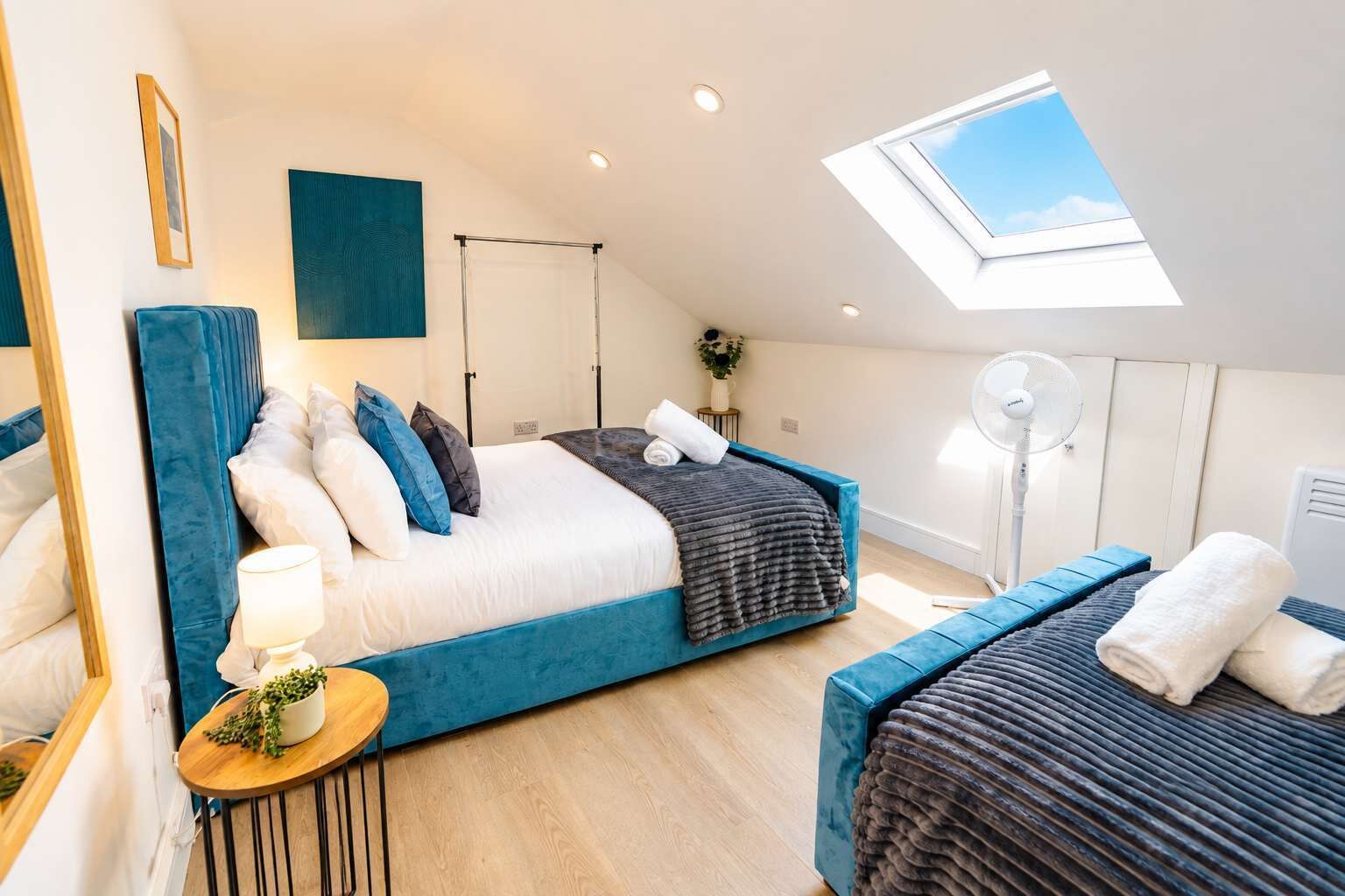 Ein modernes Schlafzimmer mit Oberlicht, einem Bett mit blauer Kopfteil, einem Nachttisch mit Lampe und einer kleinen Pflanze.