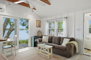 A cozy living room with a sofa, a small table, and a view of a lake through large windows. - Waterfront Cape Cottage w/ Private Beach Access (Dennis)