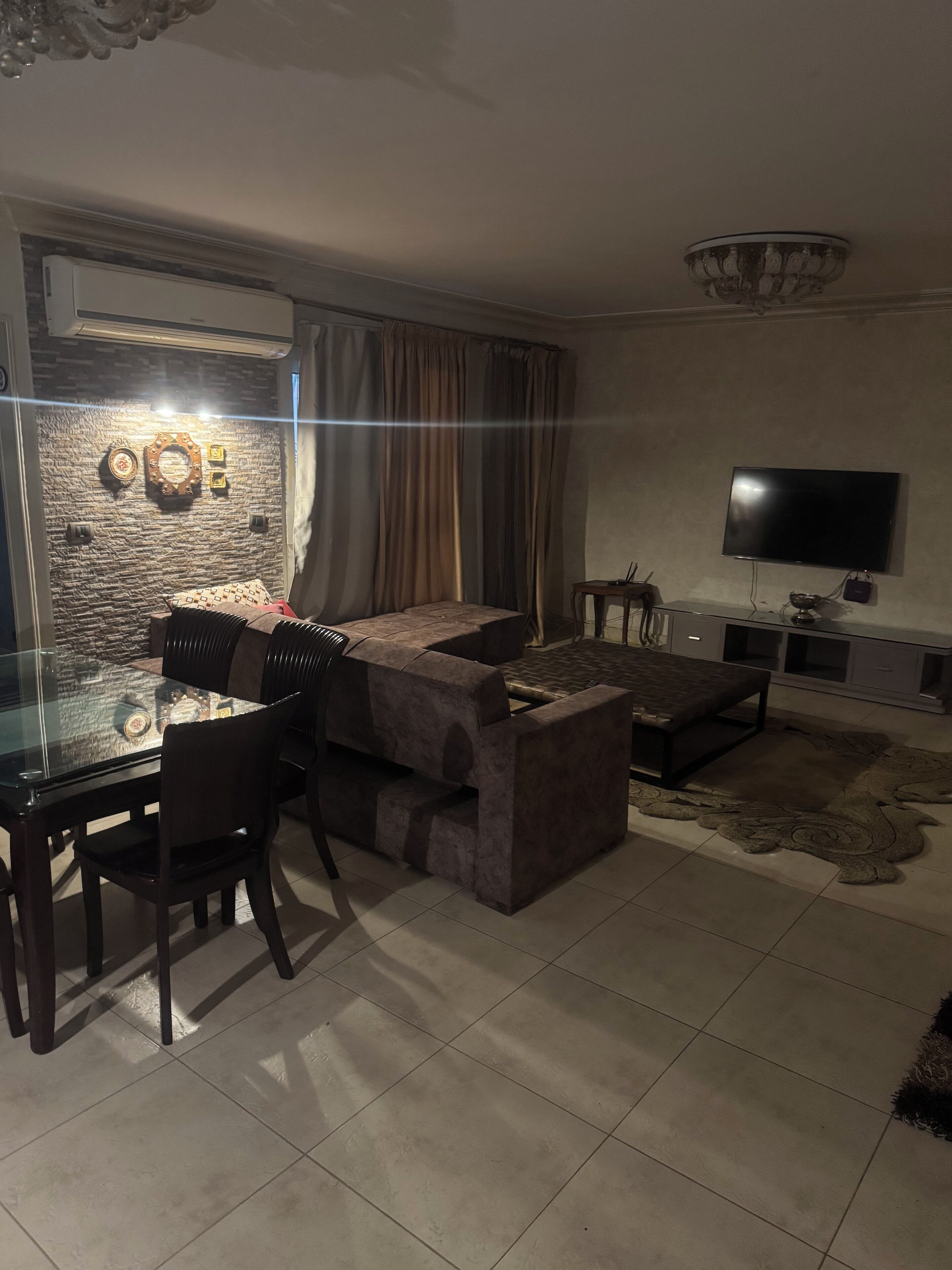 A modern living room with a glass dining table, dark sofas, a flat-screen TV, and a stone wall.