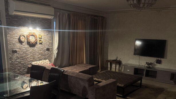 A modern living room with a glass dining table, dark sofas, a flat-screen TV, and a stone wall. - Spacious 3-bedroom apartment in charming Cairo with AC (Cairo Governorate)