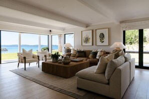 A spacious living room with a large window offering a view of the sea, a comfortable sofa set, and a coffee table. - The Farmhouse; Hauraki Views & Tranquil Luxury (Matakana)