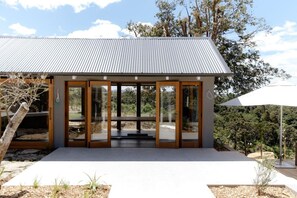 A modern building with a metal roof, large glass doors, and a patio area. - The Farmhouse; Hauraki Views & Tranquil Luxury (Matakana)