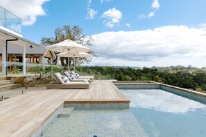 Un área de piscina moderna al aire libre con sillones de descanso y sombrillas, rodeada de una terraza de madera y vistas a un paisaje exuberante.