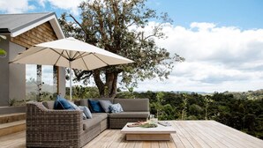 Una terraza con un sofá de mimbre, una mesa con una bebida y un árbol grande que da sombra.