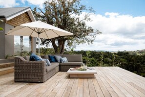 A deck with a wicker sofa, a table with a drink, and a large tree providing shade. - The Farmhouse; Hauraki Views & Tranquil Luxury (Matakana)