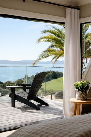 A balcony with a view of the sea, a chair, a small table with a plant, and a curtain. - The Farmhouse; Hauraki Views & Tranquil Luxury (Matakana)