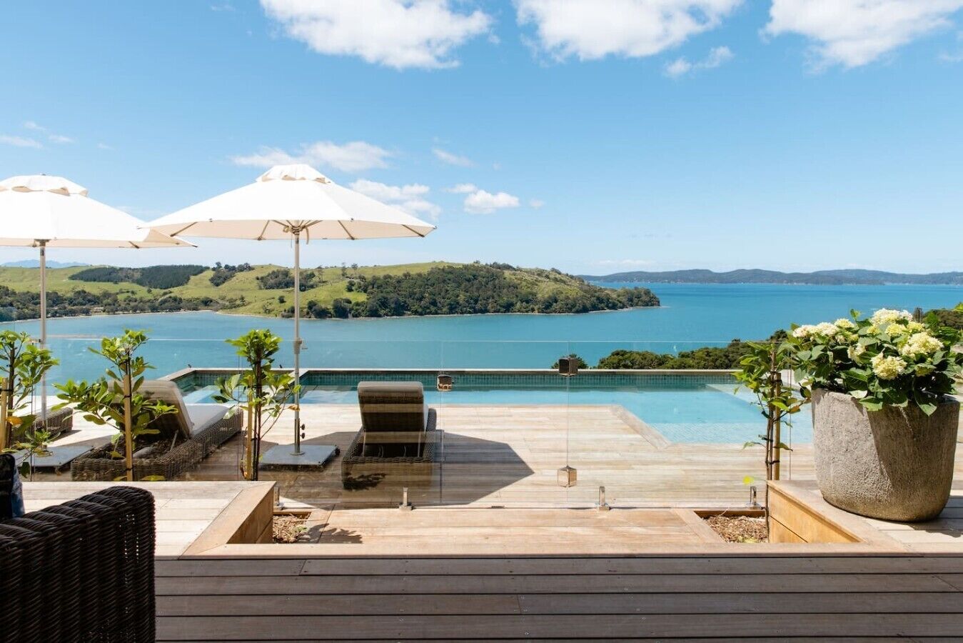 A poolside area with a view of a lake and hills, featuring lounge chairs, umbrellas, and a potted plant.