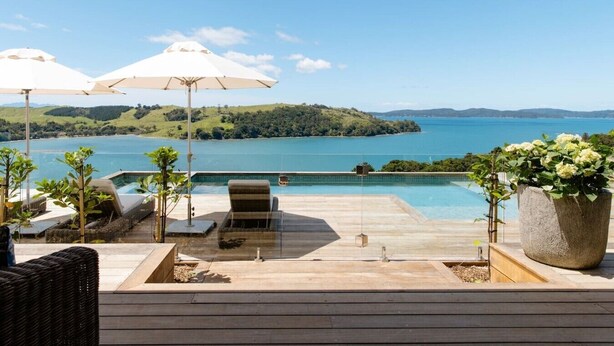 Zona junto a la piscina con vista a un lago y colinas, con sillones de descanso, sombrillas y una planta en maceta.