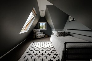 A bedroom with a sloped ceiling, a window, a bed, a chair, and a patterned rug. - The Farmhouse; Hauraki Views & Tranquil Luxury (Matakana)