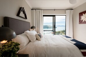 A hotel room with a large bed, a nightstand with a lamp, a view of a body of water through a glass door, and a decorative wall piece. - The Farmhouse; Hauraki Views & Tranquil Luxury (Matakana)