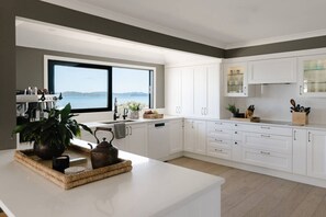 A modern kitchen with white cabinetry, a large window overlooking a body of water, and a coffee machine. - The Farmhouse; Hauraki Views & Tranquil Luxury (Matakana)