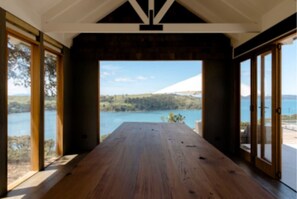 A room with a wooden floor, large windows, and a view of a lake and hills. - The Farmhouse; Hauraki Views & Tranquil Luxury (Matakana)