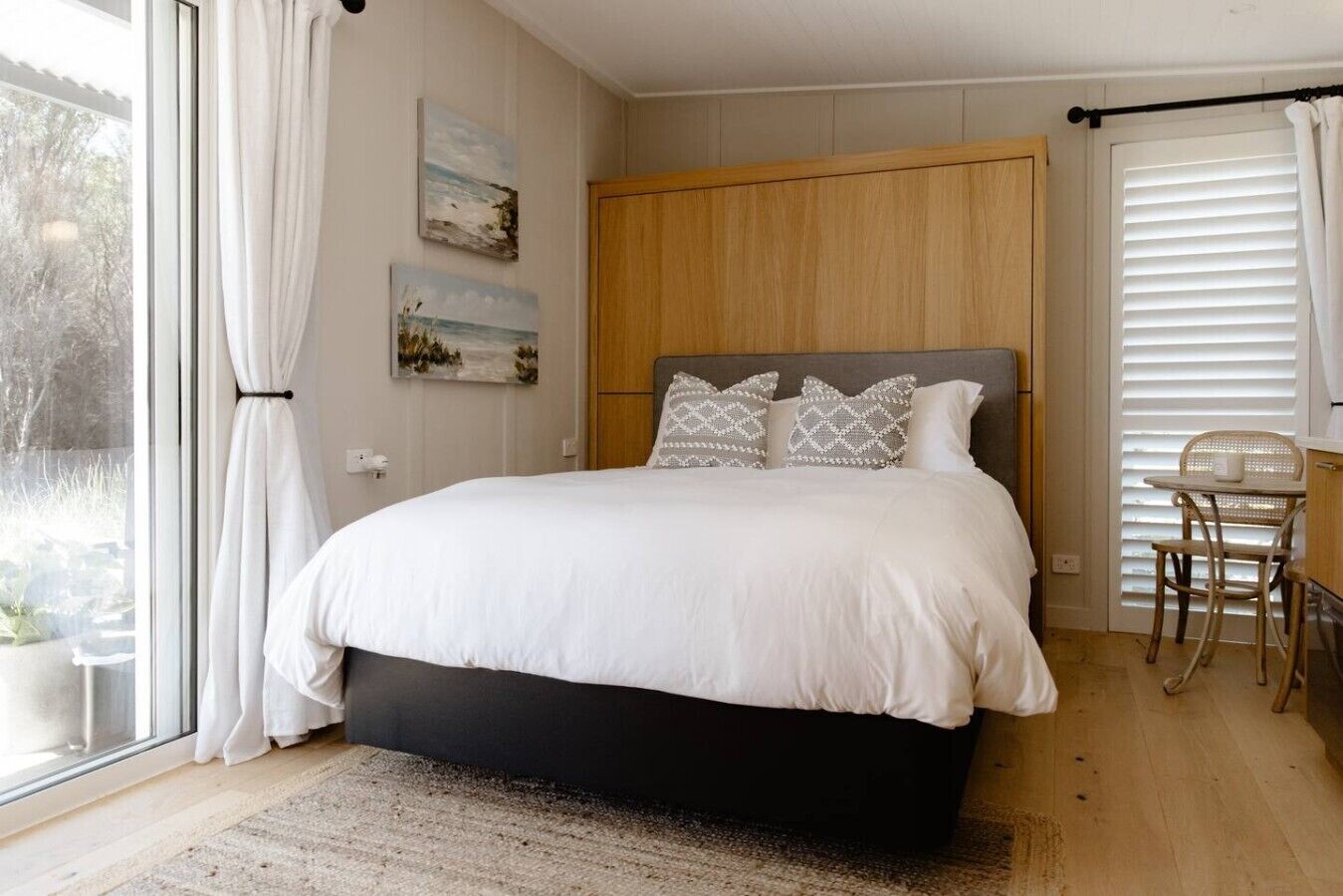 Un dormitorio con cama, ventana con cortinas, cabecero de madera, una mesita con silla y dos cuadros colgados en la pared.