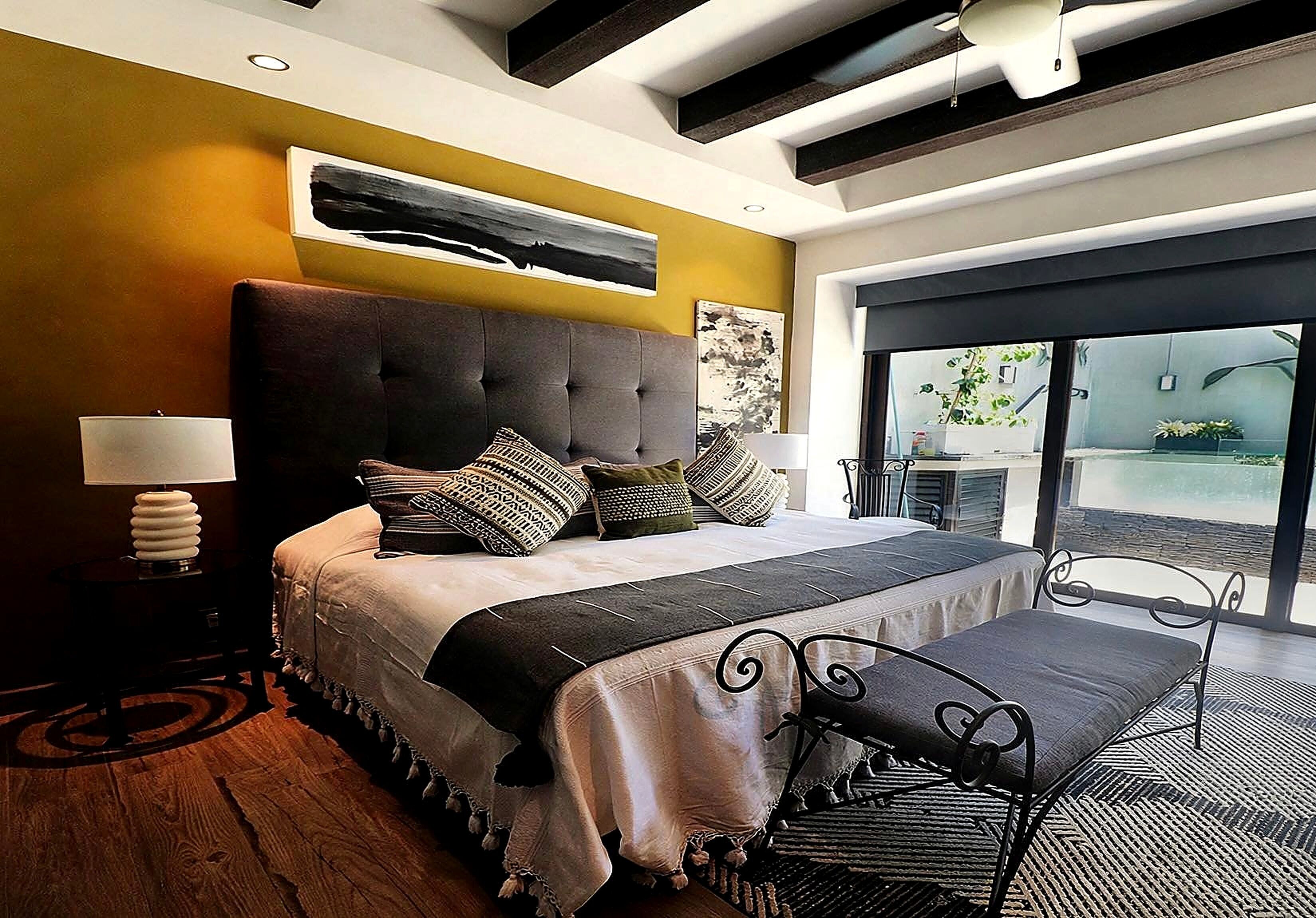 A modern bedroom with a large bed, a bench, a nightstand, and a view of the outdoors through a glass door.