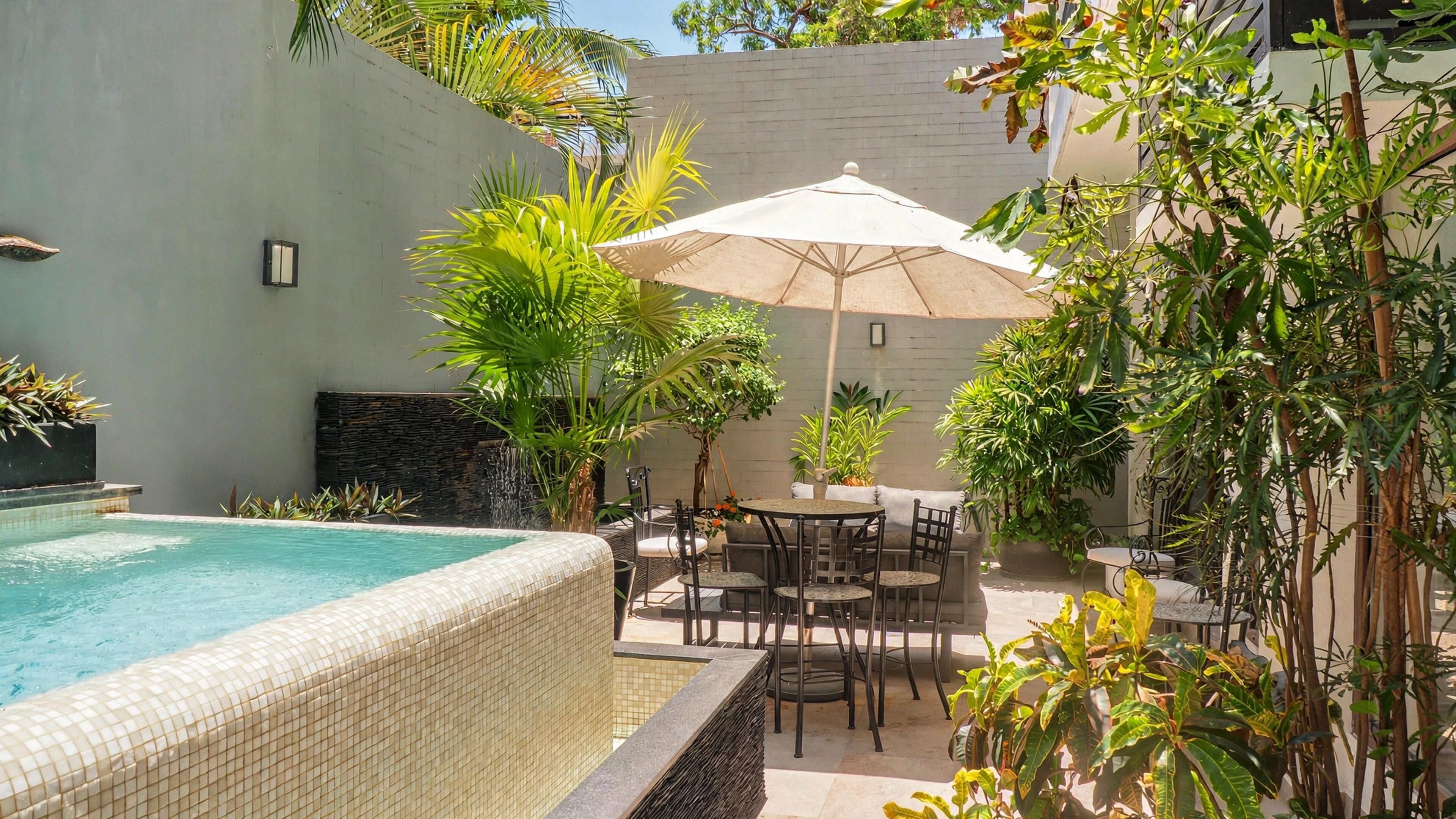 A pool with tiled edges, surrounded by a patio area with outdoor furniture and a large umbrella.