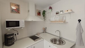 A compact kitchen with a microwave, coffee maker, stove, and sink. There are wall-mounted shelves with dishes and a potted plant. - La Alboreá (Jerez de la Frontera)