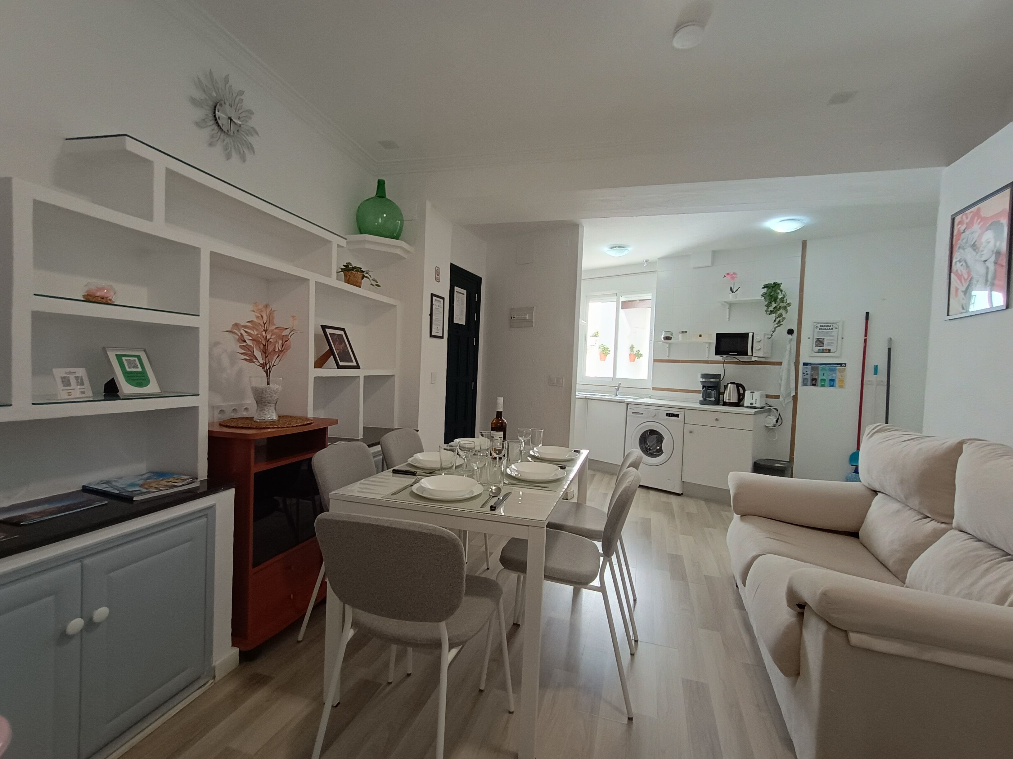 Una sala moderna con comedor, un sofá, una cocina con lavadora y una pequeña mesa de comedor lista para cuatro personas.