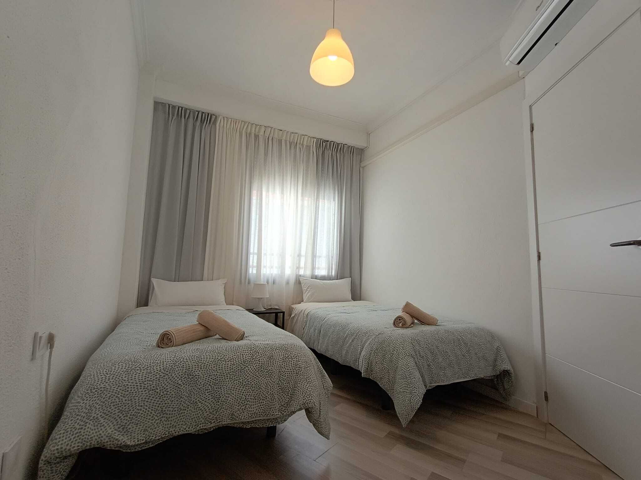 A room with two beds, a hanging light fixture, a window with curtains, and a door.