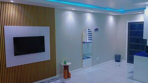 Quarto moderno com televisão de tela plana fixada em um painel de madeira, uma pequena mesa redonda com um frasco, um espelho e uma lixeira azul.