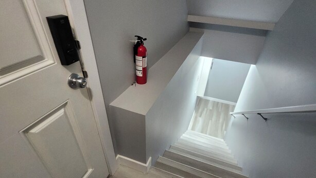 Un escalier avec un extincteur fixé au mur, une porte dotée d’un clavier pour le verrouillage, et un tapis noir à la base.