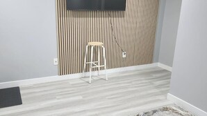 Une pièce dotée d’un téléviseur à écran plat fixé au mur, qui est recouvert de lambris verticaux, d’un tabouret blanc et d’un tapis à motifs posé sur un plancher en bois de couleur claire.