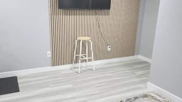Une pièce dotée d’un téléviseur à écran plat fixé au mur, qui est recouvert de lambris verticaux, d’un tabouret blanc et d’un tapis à motifs posé sur un plancher en bois de couleur claire.