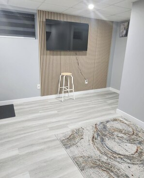 A room with a flat-screen TV mounted on a wall with vertical slats, a white stool, and a patterned rug on a light-colored wooden floor. - Sho'shaven (Winnipeg)