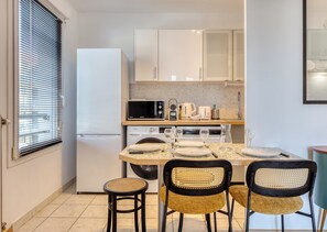 A compact kitchen with a refrigerator, microwave, and dishwasher. There is a dining table set with plates and glasses. - Floria – Élégant appartement avec balcon, cave et parking (Rouen)