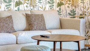 Una sala de estar con un sofá, dos almohadones estampados, una mesa de centro redonda de madera y un control remoto encima.