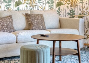 Ein Wohnzimmer mit einer Couch, zwei gemusterten Kissen, einem runden Holztisch und einer Fernbedienung darauf.