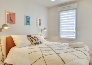 Un dormitorio con una cama, dos lámparas de noche, un ventanal con persianas y tres cuadros en la pared.