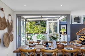 An open-plan dining area with a view of lush greenery, a wooden dining table set for six, and a staircase leading to an upper level. - Lake House by Coast Hosting (Wamberal)