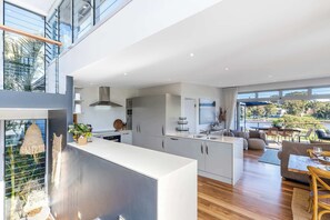 A modern kitchen with white cabinets, a central island, and a dining area with a table and chairs. - Lake House by Coast Hosting (Wamberal)