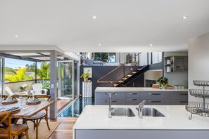 A modern kitchen with a central island, a dining area with a wooden table, and a staircase leading to an upper level. - Lakefront. Terrigal Beach Walk. Sleeps 10 (Wamberal)