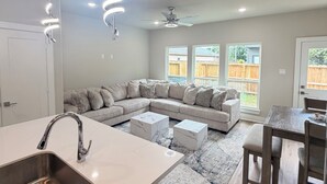 A modern kitchen with a built-in sink, a dining area with a wooden table and chairs, a large L-shaped sectional sofa, and a ceiling fan. - Houston Hub- Family Getaway, Garage & Yard (Houston)
