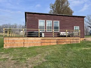 Une cabane en bois dotée d’une terrasse entourée d’une clôture, d’un petit porche et d’une façade avec des fenêtres.