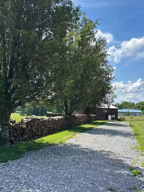 Un chemin de gravier mène à un bâtiment rustique doté d’un mur en pierre et d’un grand arbre qui offre de l’ombre.