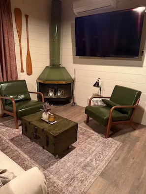 A cozy room with a fireplace, two green leather chairs, a small table with a vase, and a flat-screen TV mounted on the wall.
