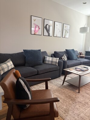 Ein modernes Wohnzimmer mit einem grauen Sofa, einem braunen Ledersessel und einem Couchtisch mit Büchern und einer Fernbedienung.