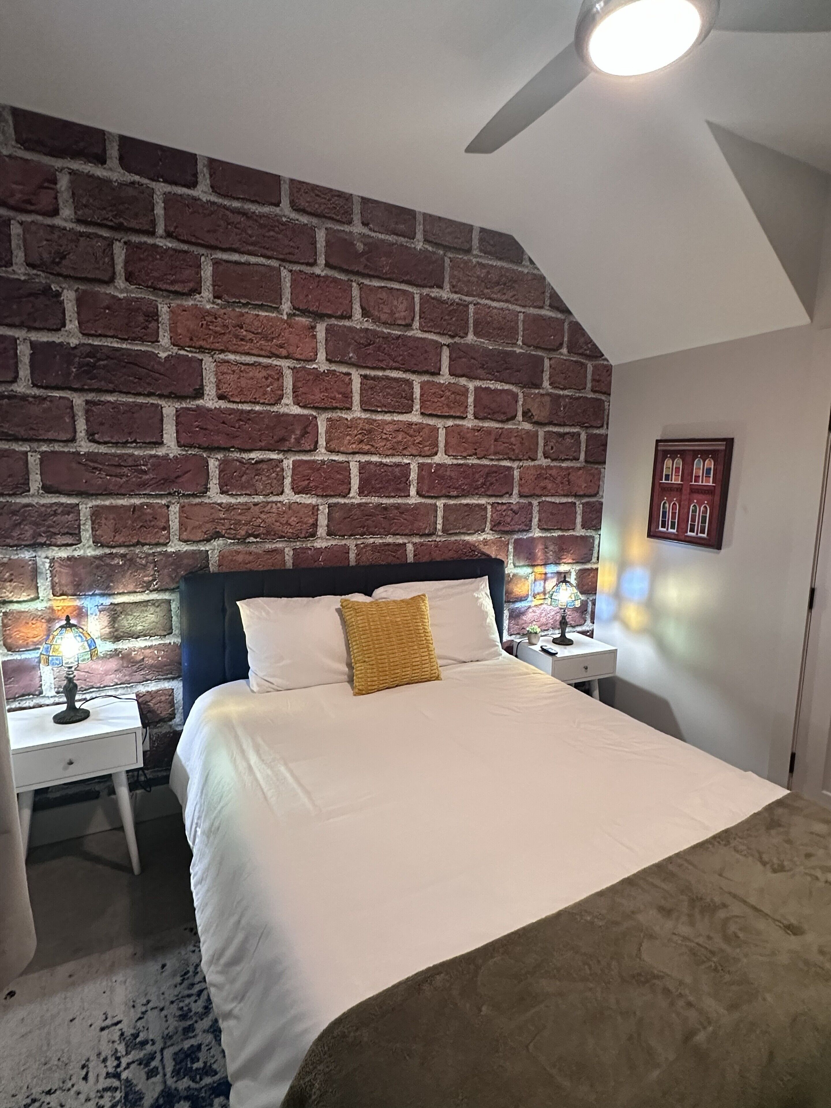 A bedroom with a brick wall, a bed with white bedding, two bedside tables with lamps, and a ceiling fan.